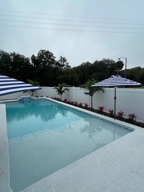 Pool with striped umbrellas, palm trees, and white wall in a residential setting.
