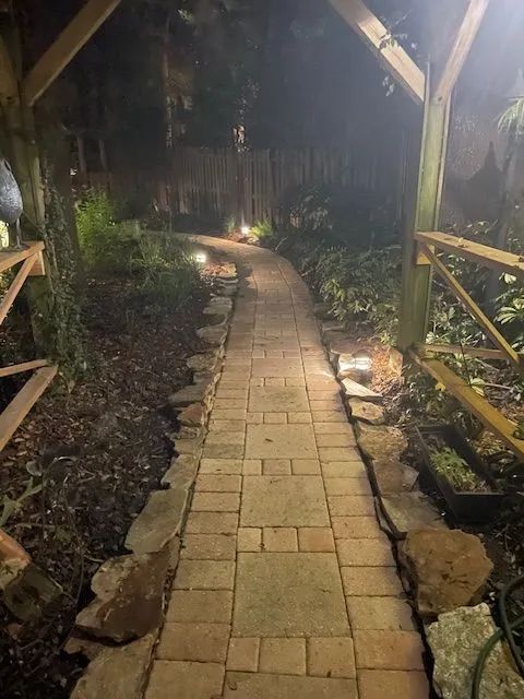 Brick pathway through a garden at night, illuminated by pathway lights under a wooden arbor.