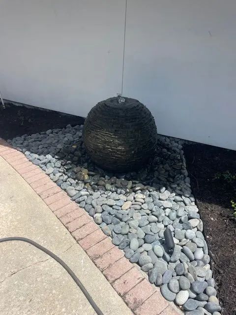Spherical water fountain on a bed of river rocks next to a sidewalk and a white wall.