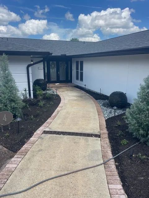 Sidewalk leading to a white house with black trim. Landscaping lines the path with greenery and mulch.