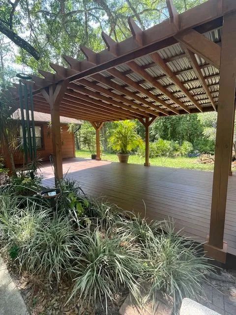 Wooden pergola over a deck, surrounded by plants and trees, with a house in the background.