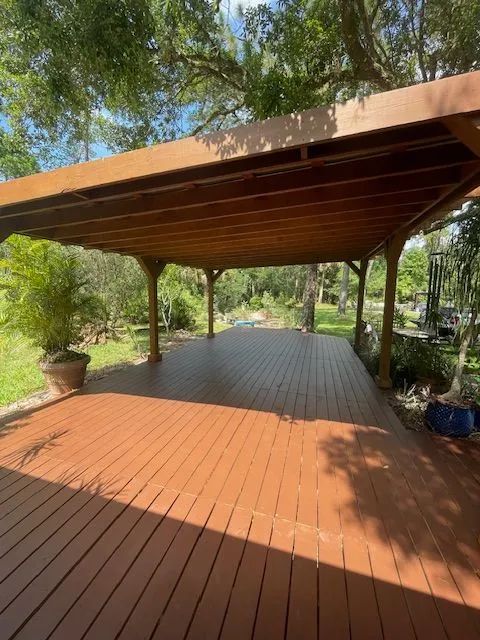 Wooden deck with brown stain, under a covered patio with trees in the background.