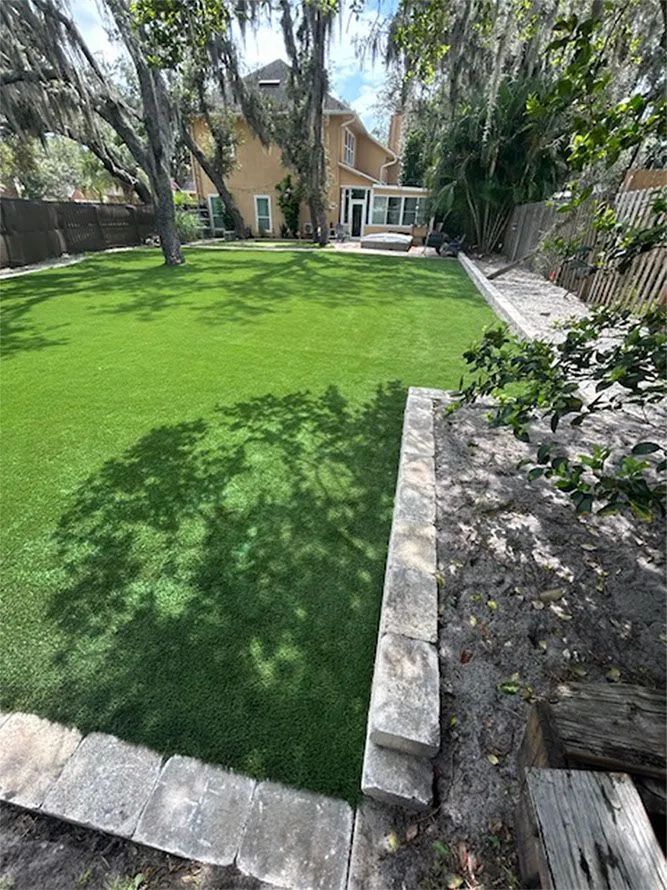 Backyard with artificial green grass, stone retaining wall, and house in the background.