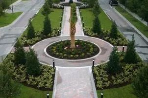Landscaped roundabout with pathways and trees, surrounded by roads, in a sunny setting.