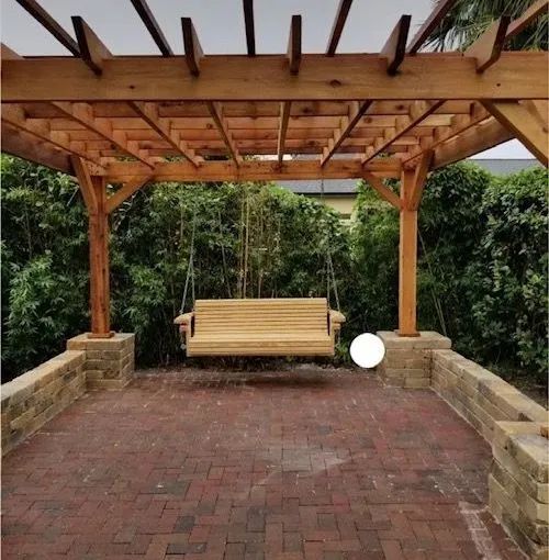 Wooden swing hanging under a wooden pergola on a brick patio.