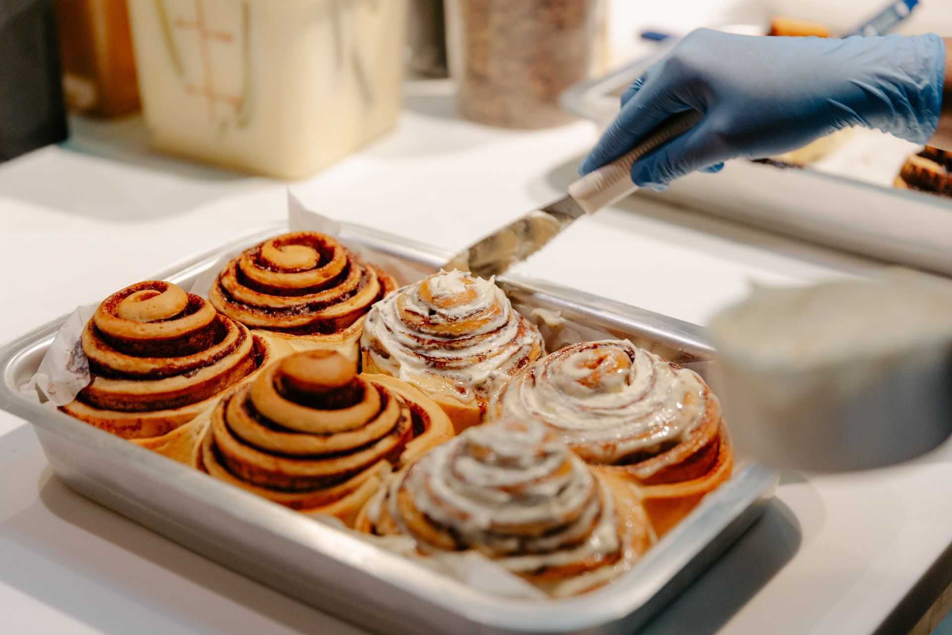Gloved hand arranging cinnamon rolls with frosting on a tray in a bakery kitchen