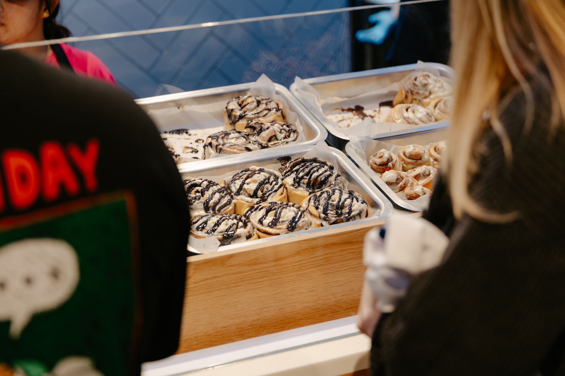 Trays of chocolate-drizzled Cinnabon at a market stall, with people browsing nearby