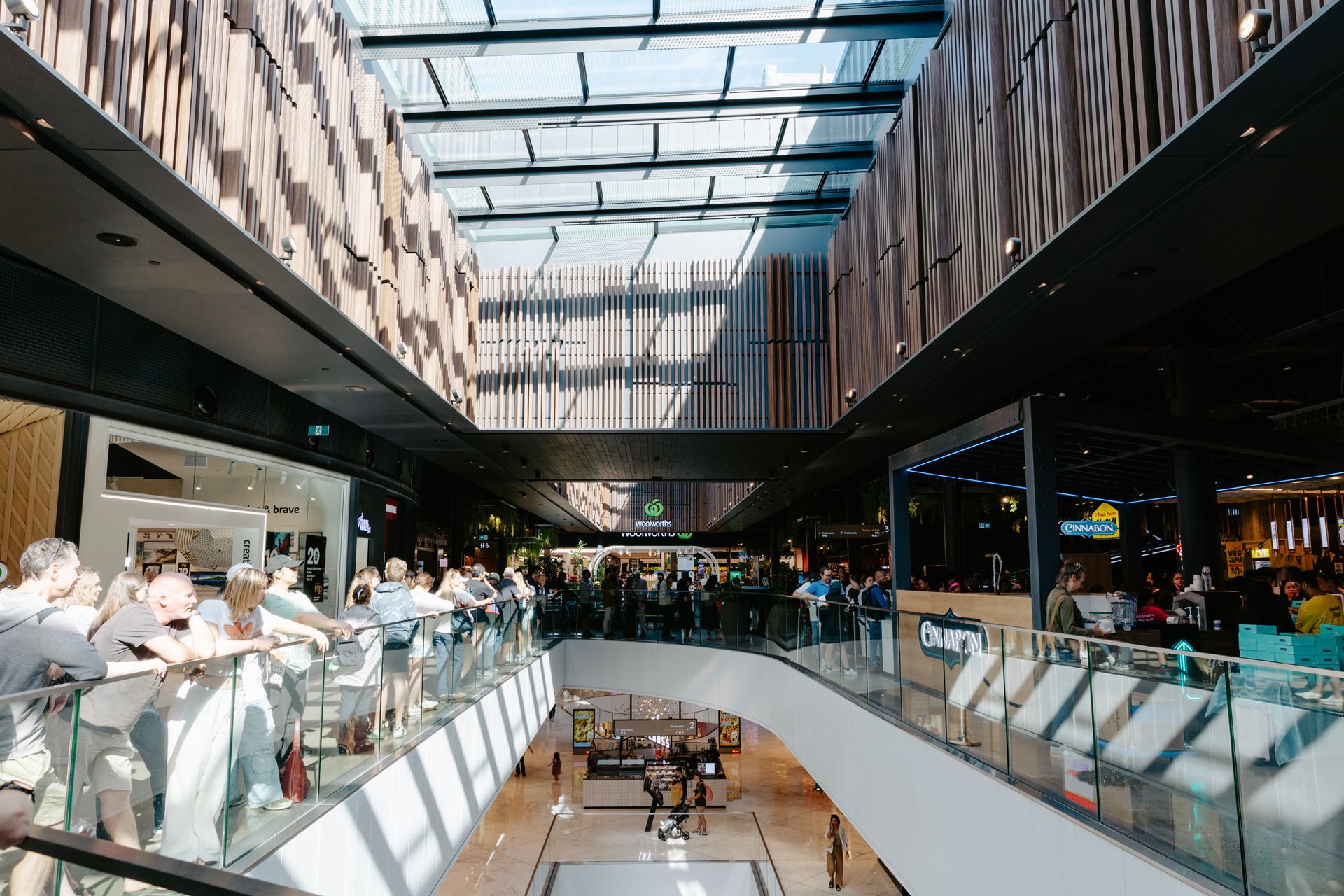Modern shopping mall atrium with skylight, glass railings, and people seated at tables on multiple levels