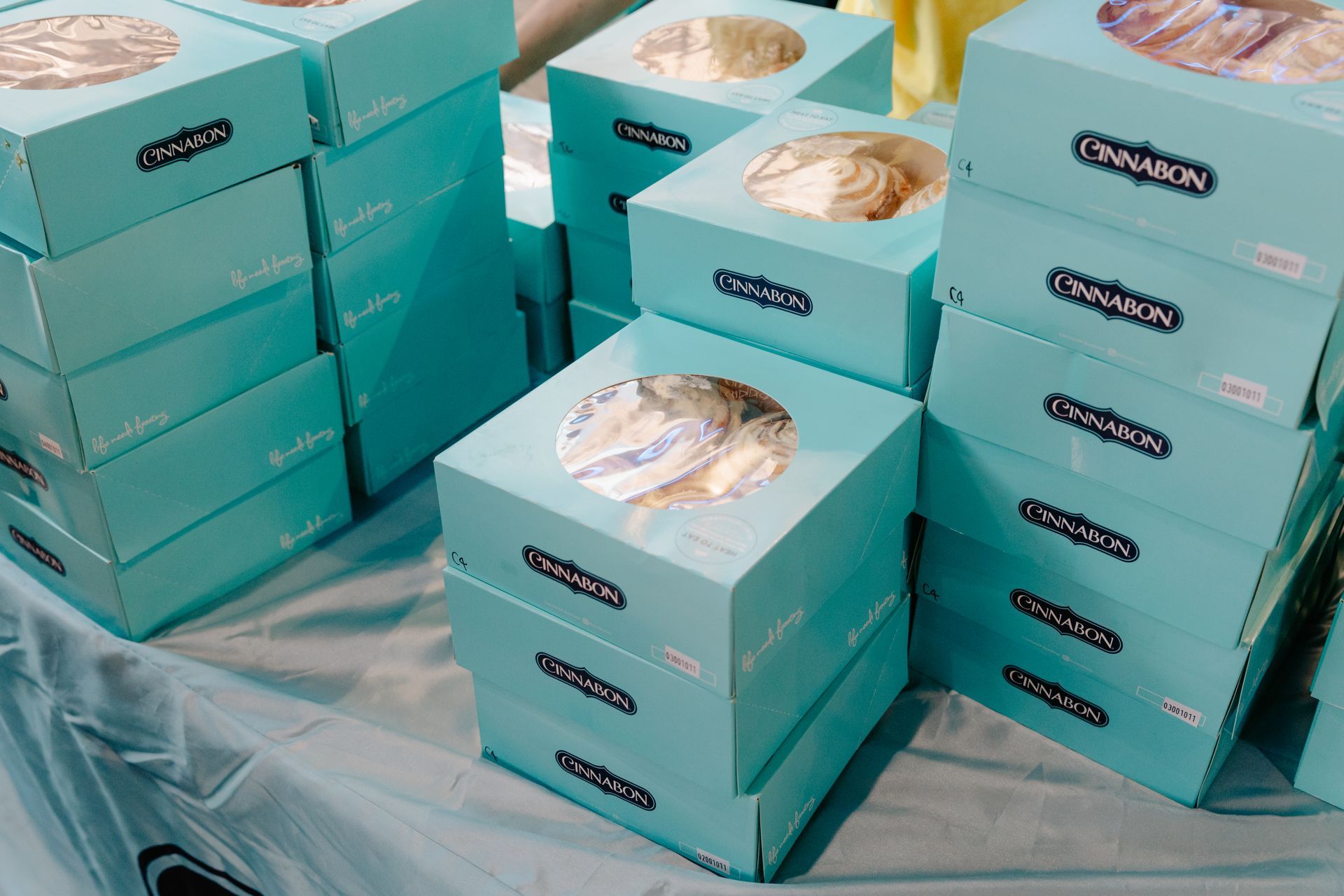 Stacks of turquoise Cinnabon gift boxes with clear lids, arranged on a table covered in plastic.