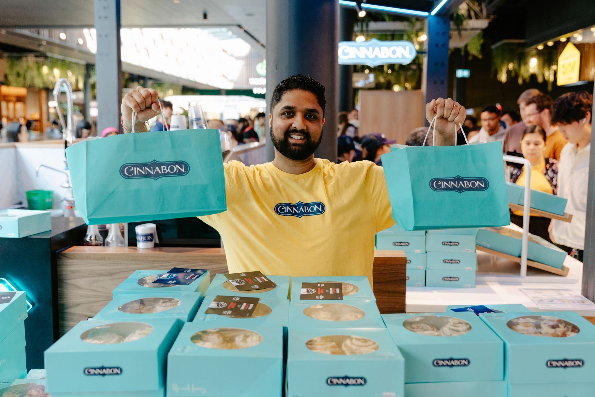 Man in yellow shirt selling turquoise Cinnabon boxes at a crowded bakery stand