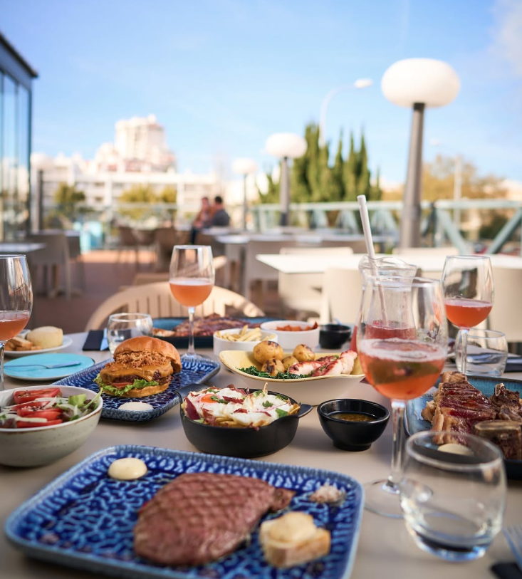Mesa de jantar ao ar livre no terraço com bifes, hambúrgueres, bebidas e vista da cidade ao pôr do sol.