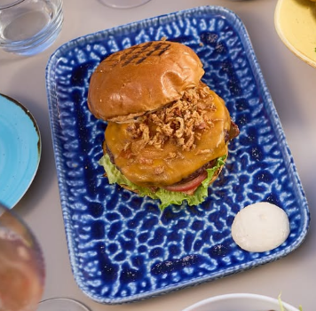 Hambúrguer com cebola frita em um prato azul estampado com uma porção de molho branco.