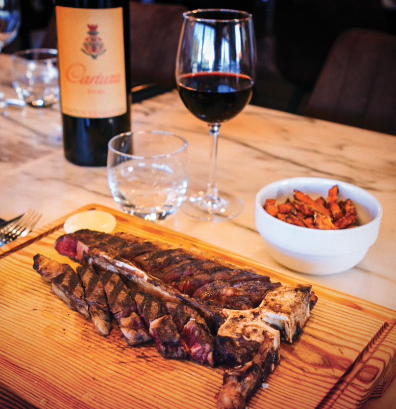 Bife grelhado em tábua de madeira com uma taça de vinho tinto, uma garrafa de água e uma tigela de acompanhamentos.