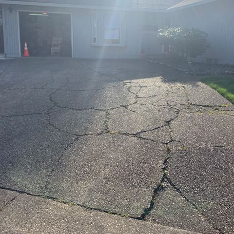Cracked asphalt driveway in front of a house, with sunlight glare and a traffic cone near the garage.