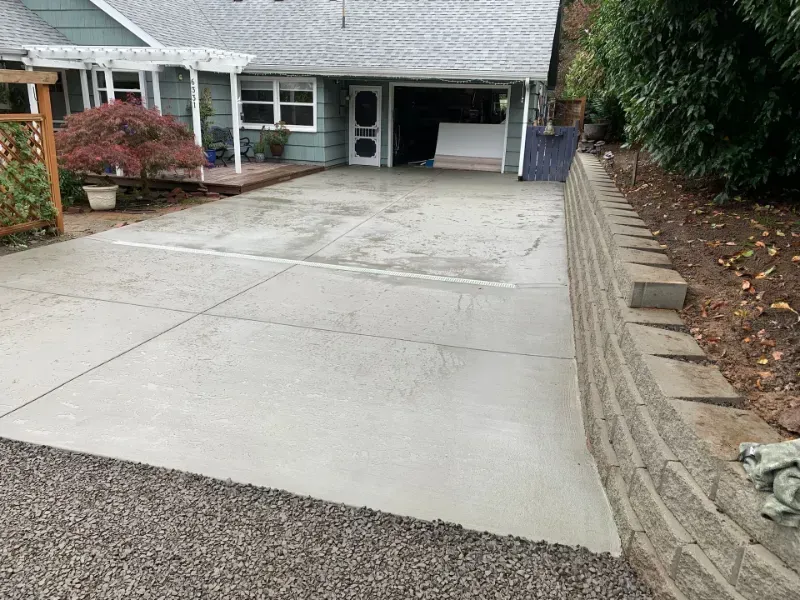 A newly poured gray concrete driveway leads to a garage, bordered by a tiered stone retaining wall on the right.