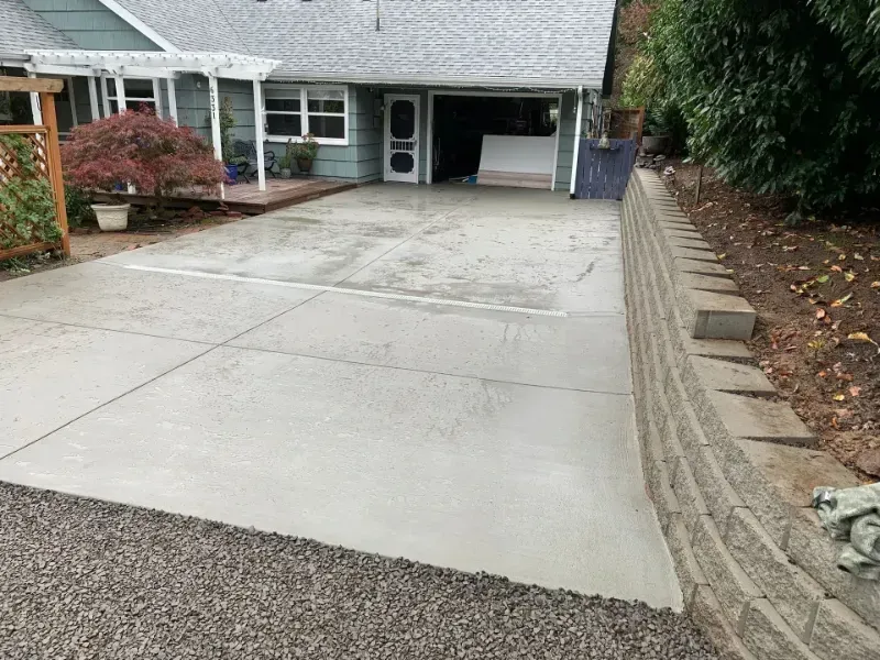 Newly poured concrete driveway in front of a blue house with a retaining wall on the right.