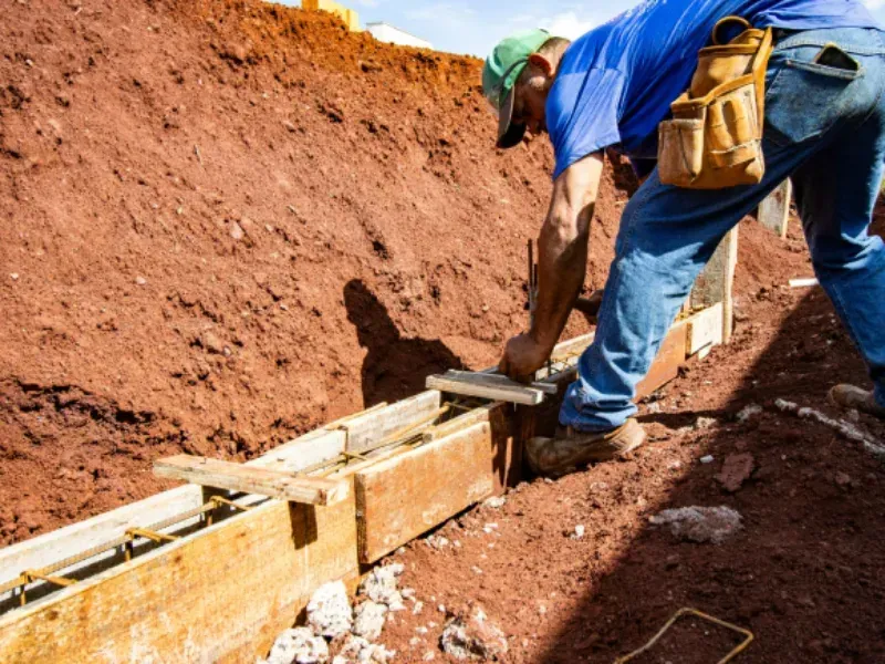 Man in blue shirt and jeans constructing a wooden form on a construction site, next to red soil.