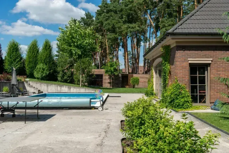 Backyard patio with an above-ground pool, green shrubs, and a brick house under a sunny blue sky