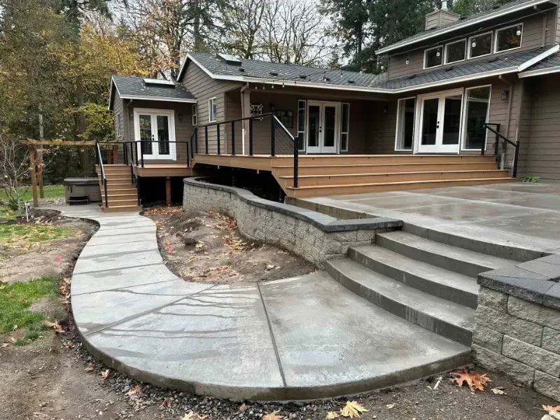 Curving concrete pathway leading to a house with a raised wooden deck and steps. Brown siding, black railing.