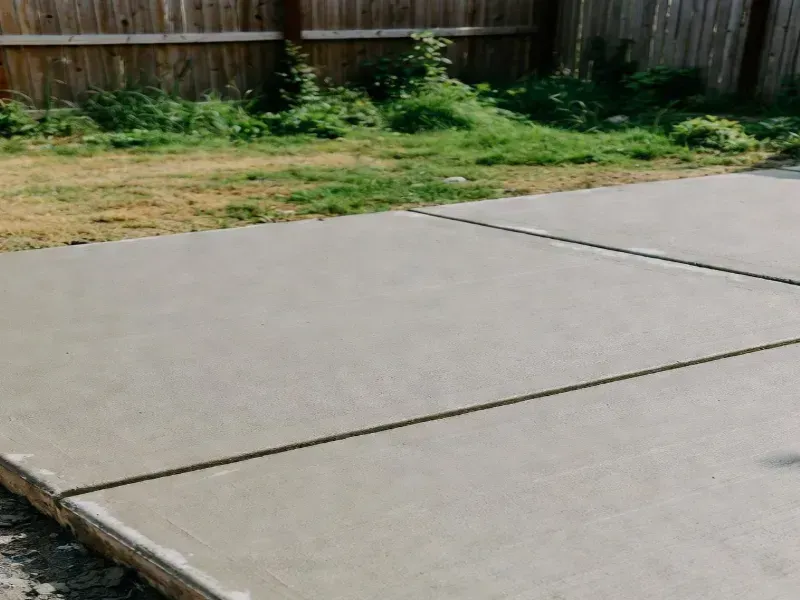 Freshly poured concrete patio with dark lines, adjacent to a grassy yard and wooden fence.