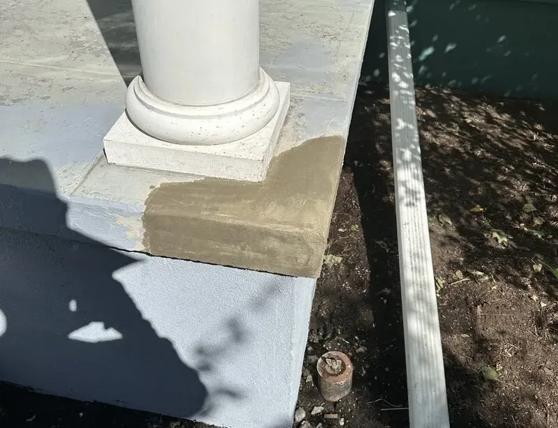 Concrete porch corner with wet, light-brown patch near a white column base; a downspout and dirt are in the background.
