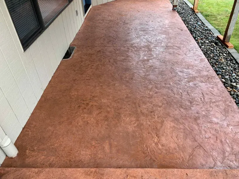 Brown stamped concrete patio next to a white house with a rock border.