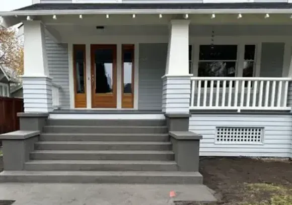 A Craftsman-style house porch with gray stairs, white tapered pillars, gray siding, and a wooden front door.