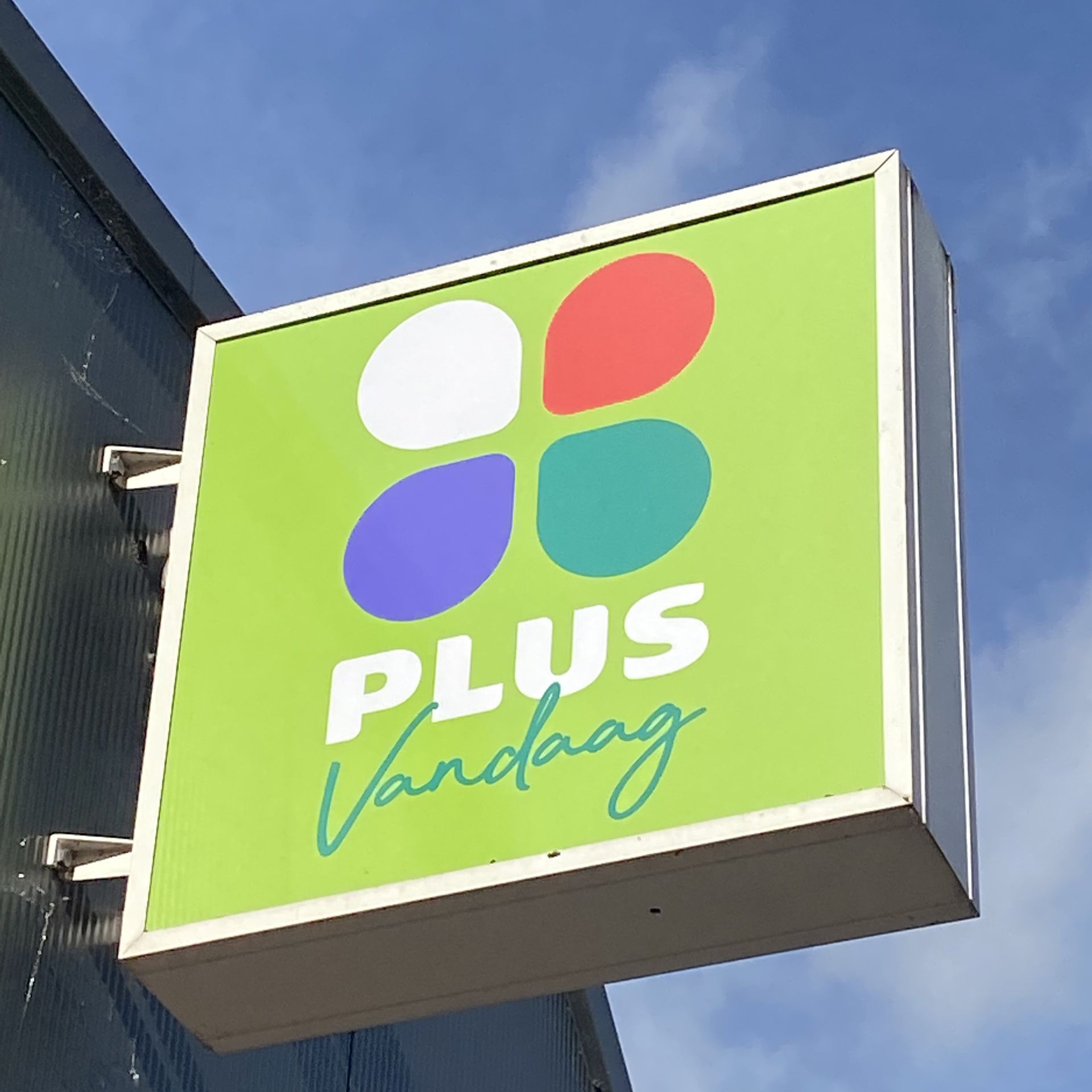 Winkelgevelbord van Green Plus Värmdag met vier kleurrijke bloemblaadjes tegen een blauwe lucht.