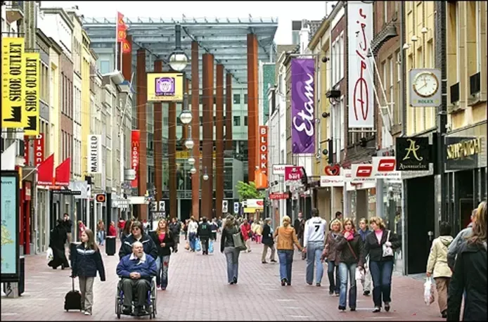 Een drukke winkelstraat voor voetgangers met etalages, uithangborden en mensen die voorbijlopen.