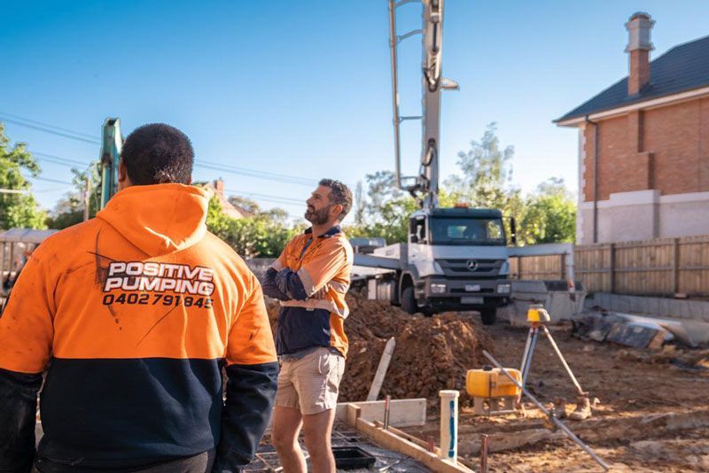 Two construction workers are standing in front of a cement pump on a construction site.