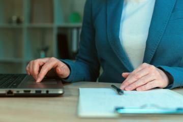 A person in a blue blazer types on a laptop next to a notepad and pen on a desk in a professional office.