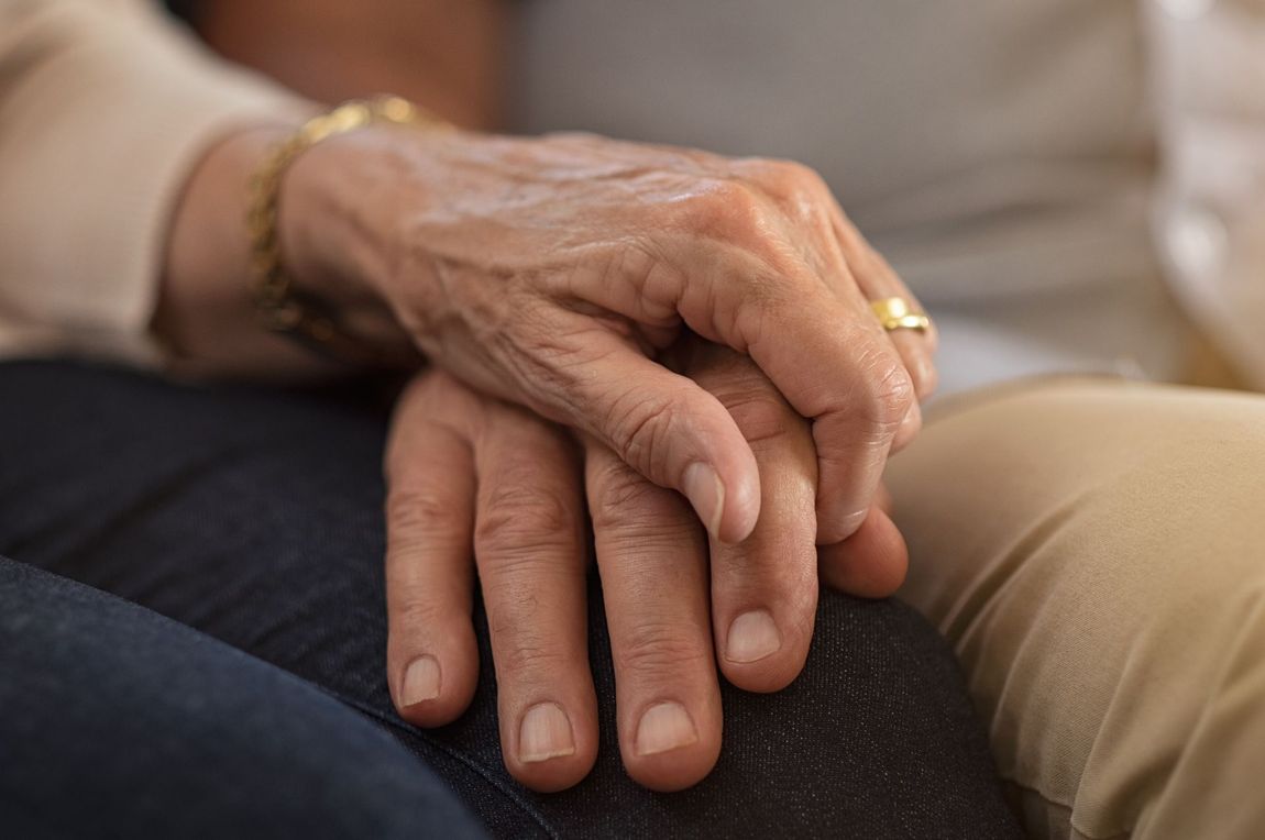 An elderly couple is holding hands while sitting on a couch.