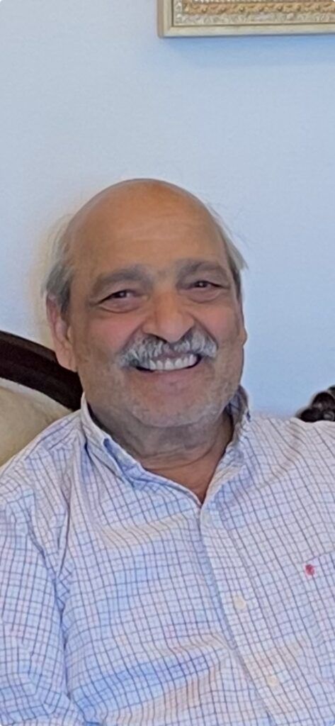 Smiling man in a light checkered shirt sitting indoors against a pale blue wall.