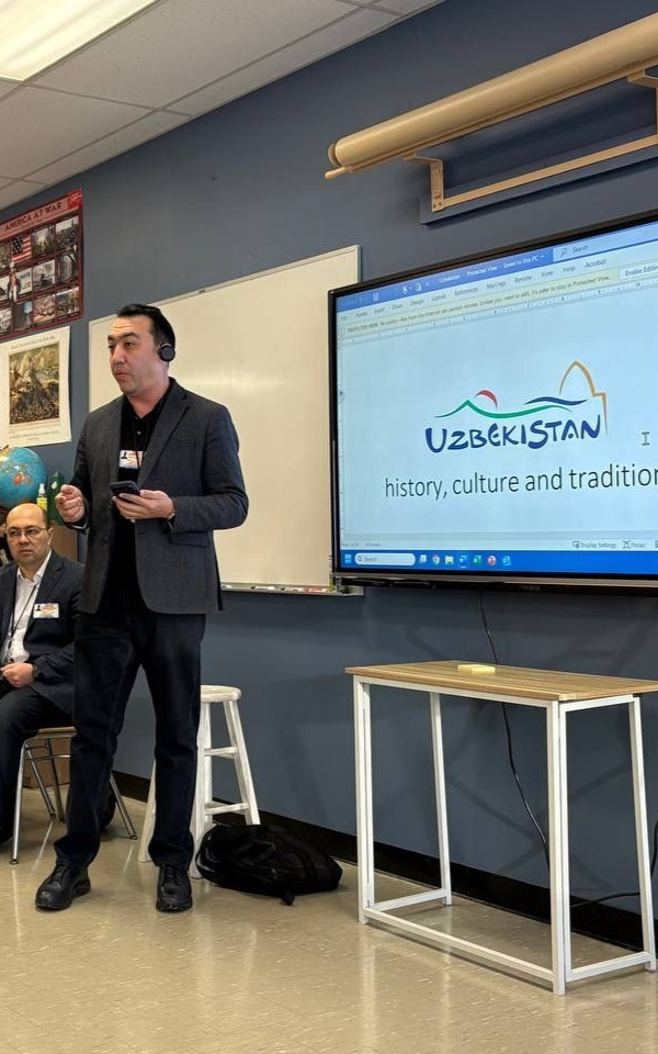 Man presenting beside a classroom display with a slide titled “Uzbekistan.”