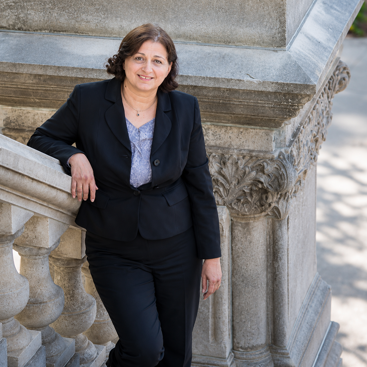 Woman in a black suit leaning on a stone balustrade outdoors, smiling at the camera.