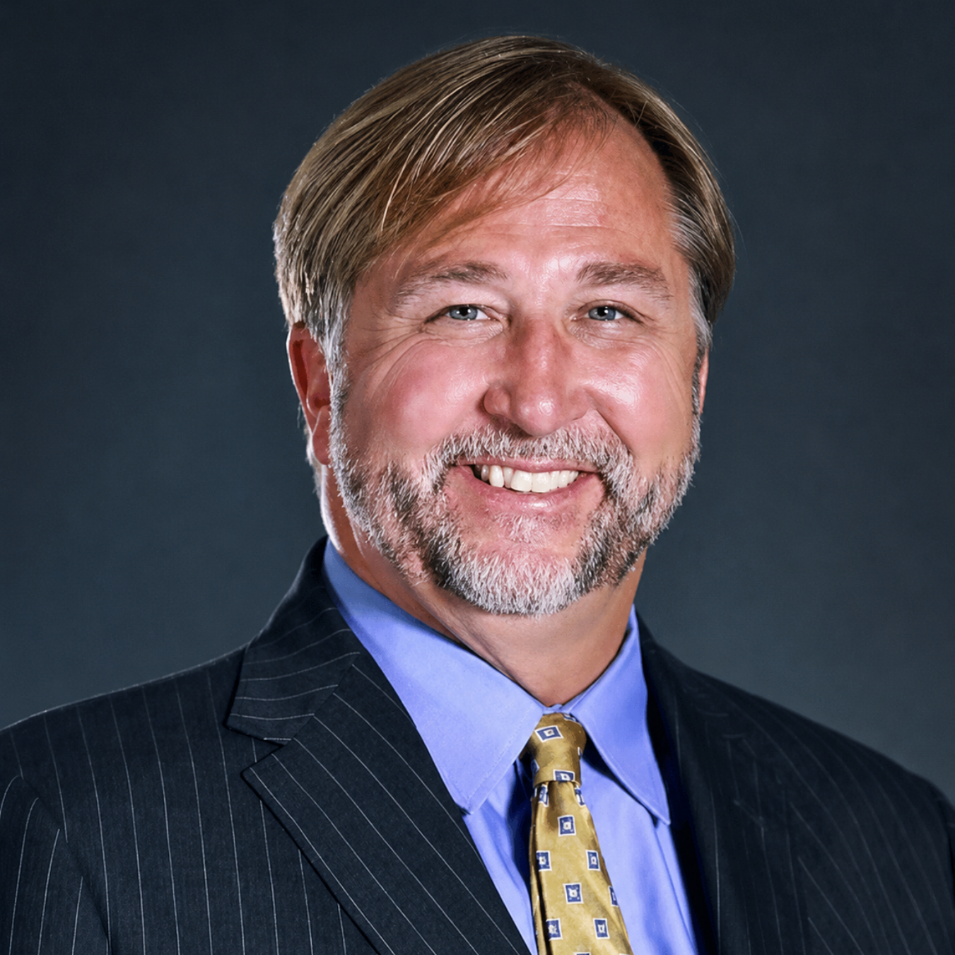 Studio portrait of a smiling man in a dark pinstripe suit and blue shirt against a gray background