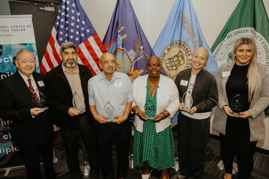 Six people standing in a row at a formal event with U.S. and organizational flags behind them