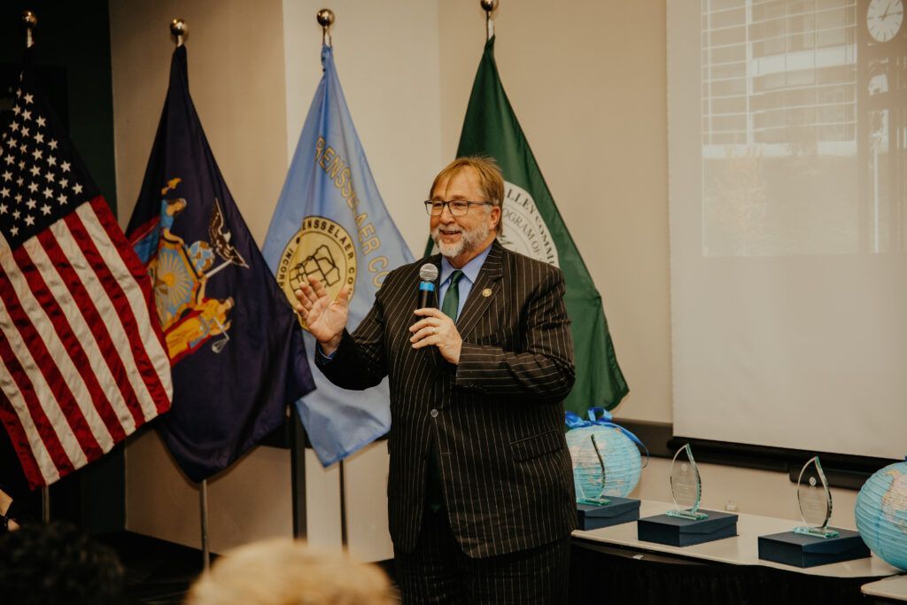 Speaker holding a microphone at a podium beside U.S. and state flags in a meeting room