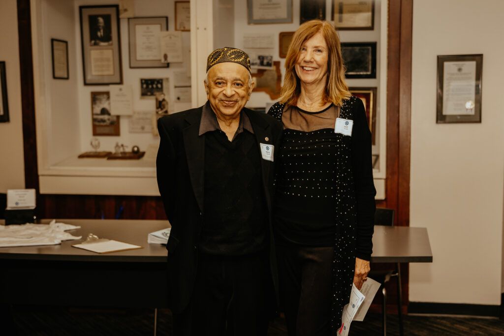 Two people standing indoors in front of framed photos, smiling at the camera.