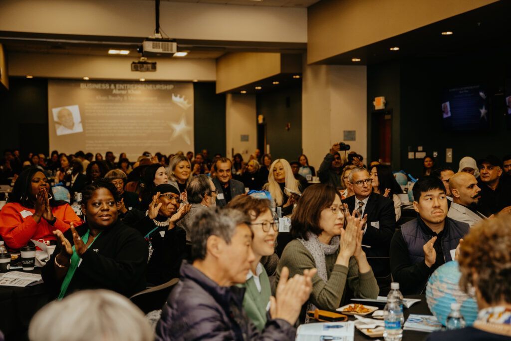 Audience seated at tables in a conference hall, listening attentively during a presentation