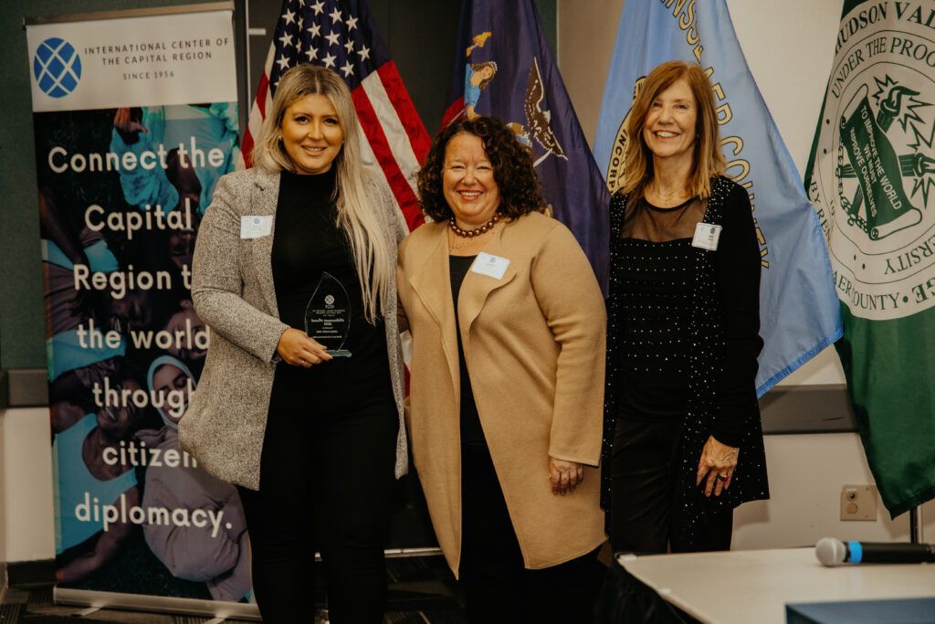 Three women pose indoors beside banners and an American flag at a civic event.