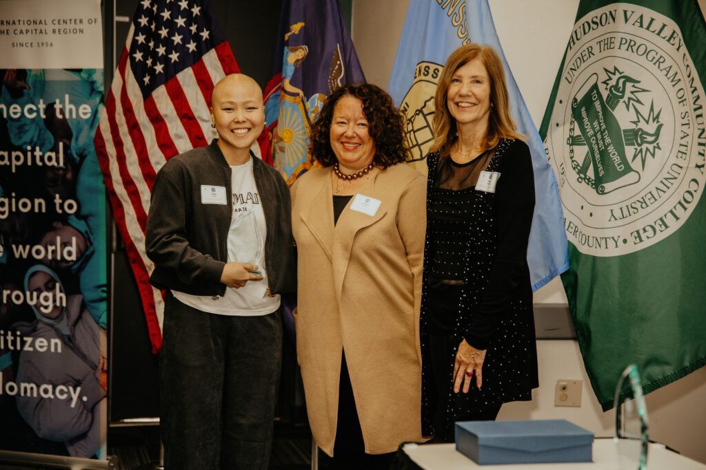 Three people posing at an event booth with flags and a banner in the background.