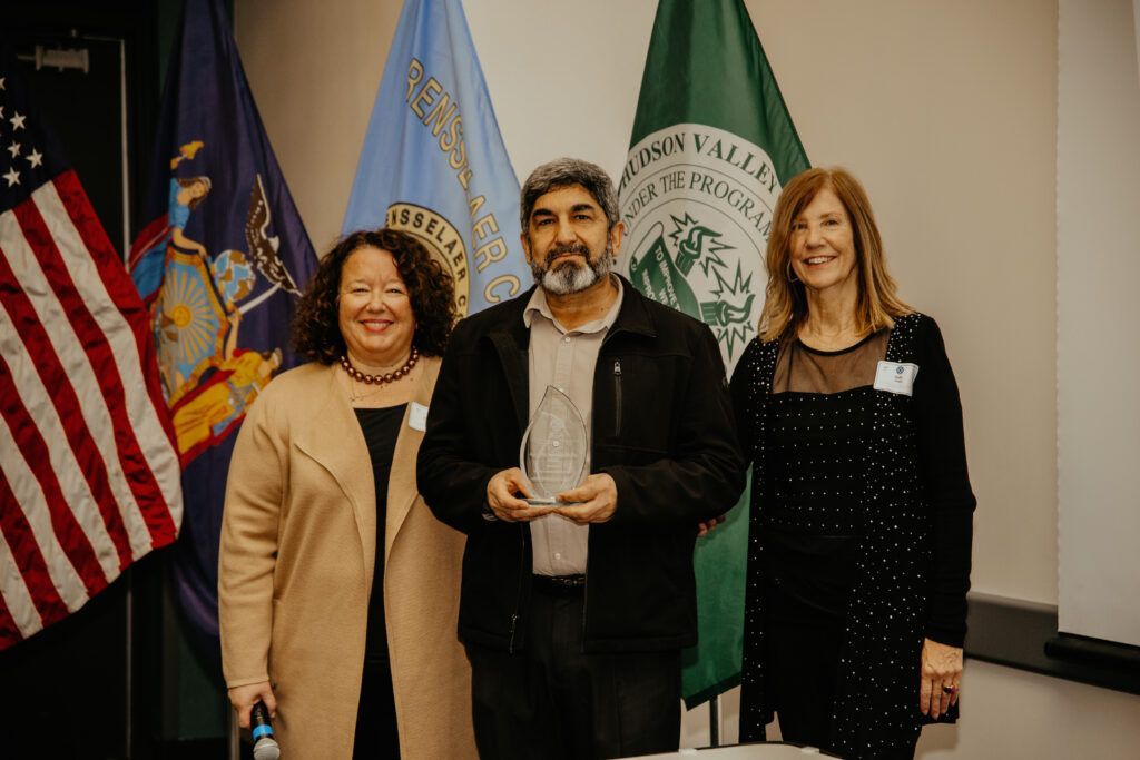 Three people posing indoors in front of flags, one holding a small award.