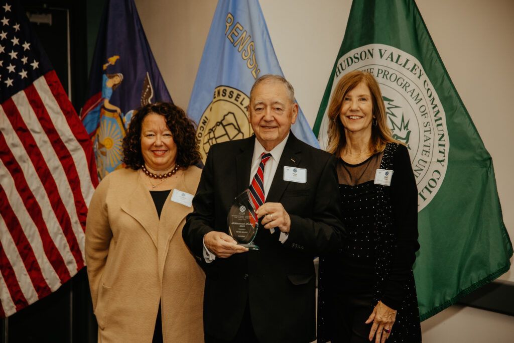 Three people posing with an award in front of flags at a ceremony