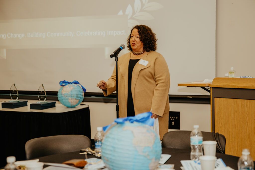 Woman speaking at a conference table with blue globe decorations and a microphone in front of a projection screen