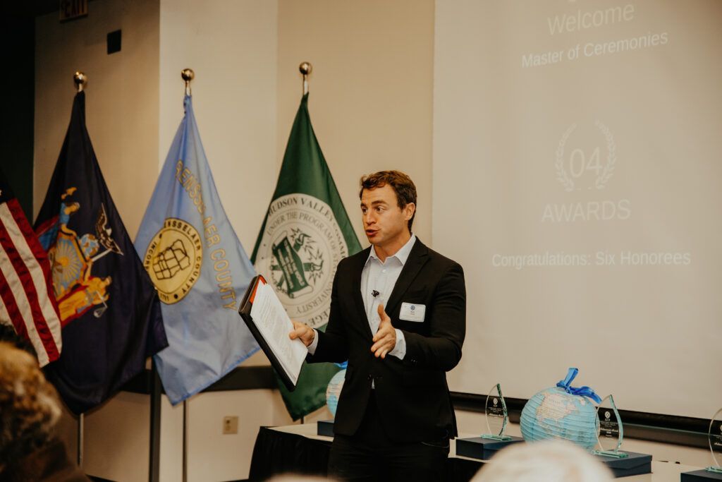 Man speaking beside flags in a conference room, holding a folder and gesturing during a presentation.