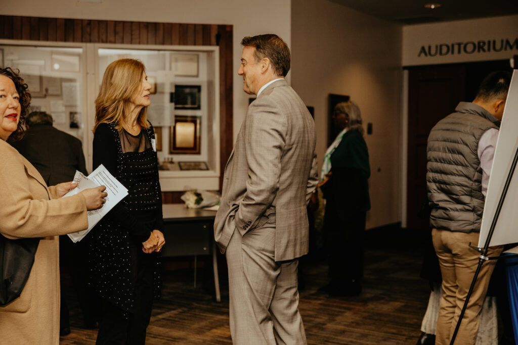 People conversing in a hallway outside an auditorium, with one man in a gray suit and others holding papers.
