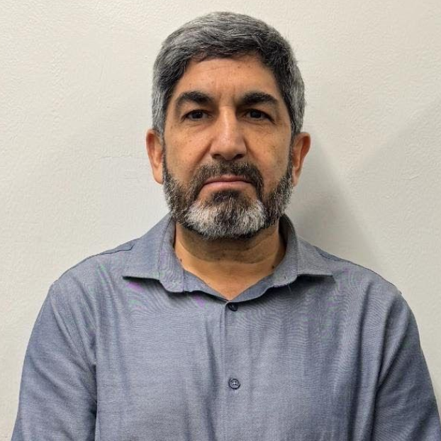 Man with salt-and-pepper beard in a gray polo shirt, standing against a plain light wall.