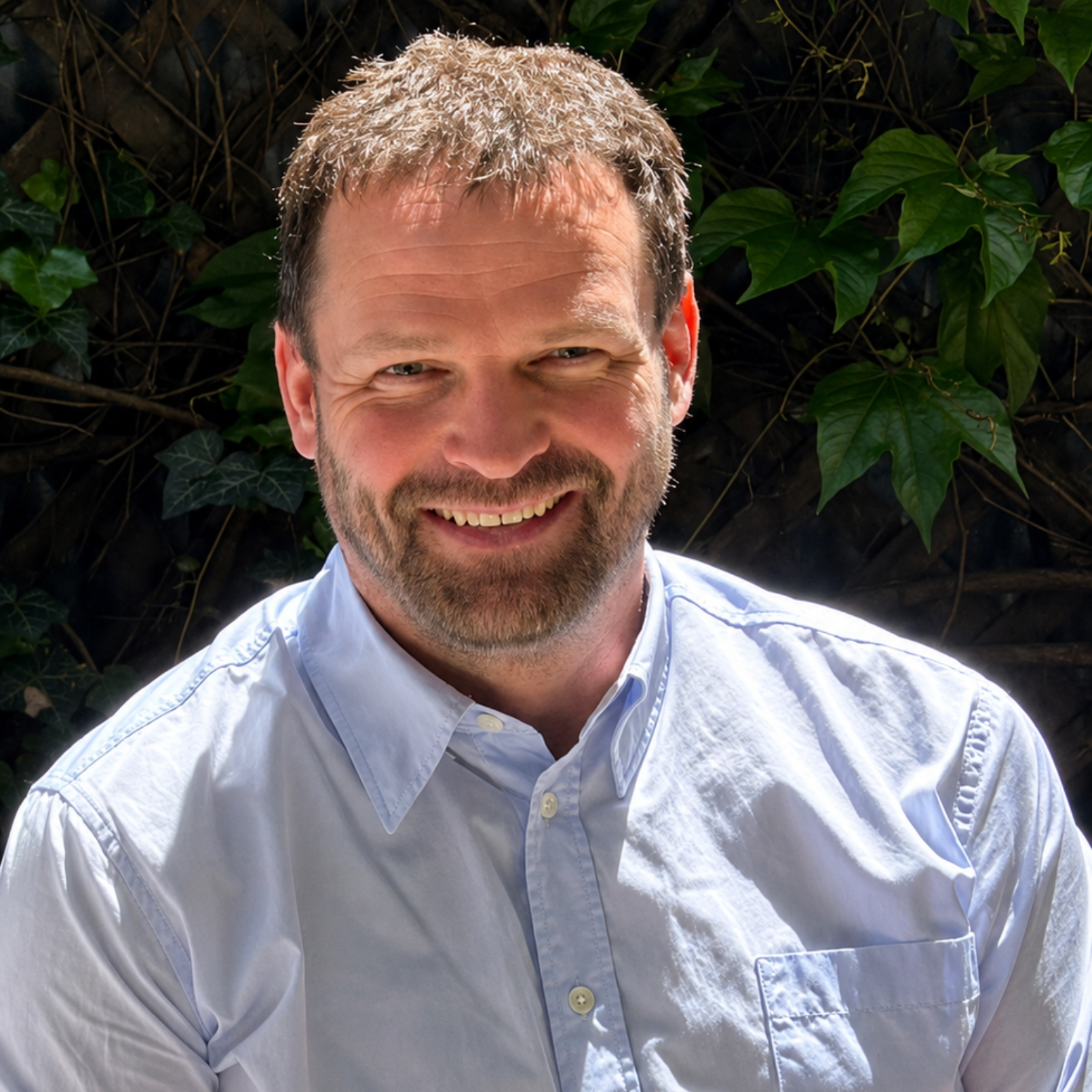 Smiling man in a light blue shirt standing outdoors in front of leafy greenery