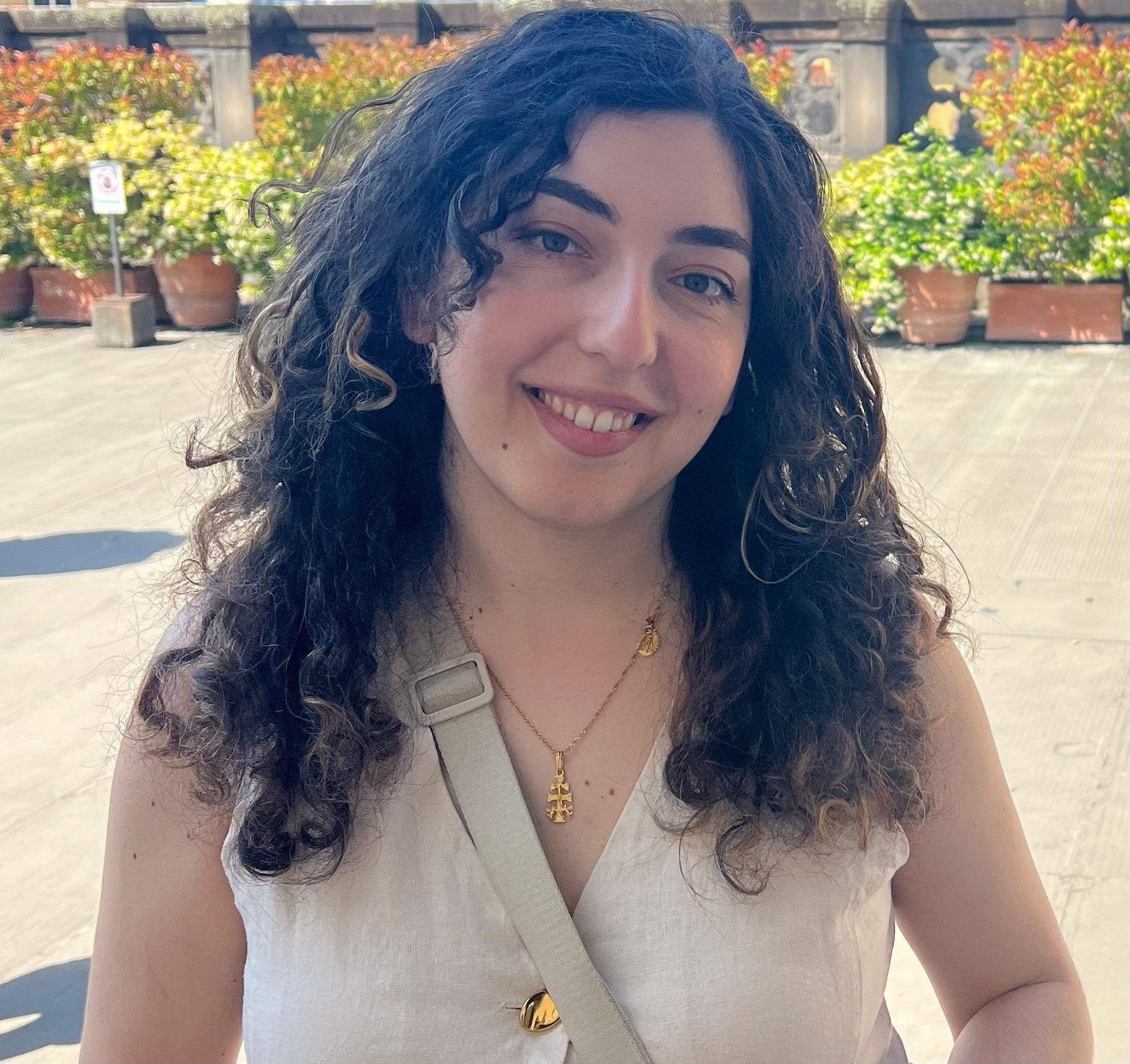 Smiling person with curly hair in a cream top, standing outdoors with potted plants in the background